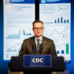 An anonymous spokesperson stands at a podium in a briefing setting with an infographic backdrop, conveying clarity and professionalism.