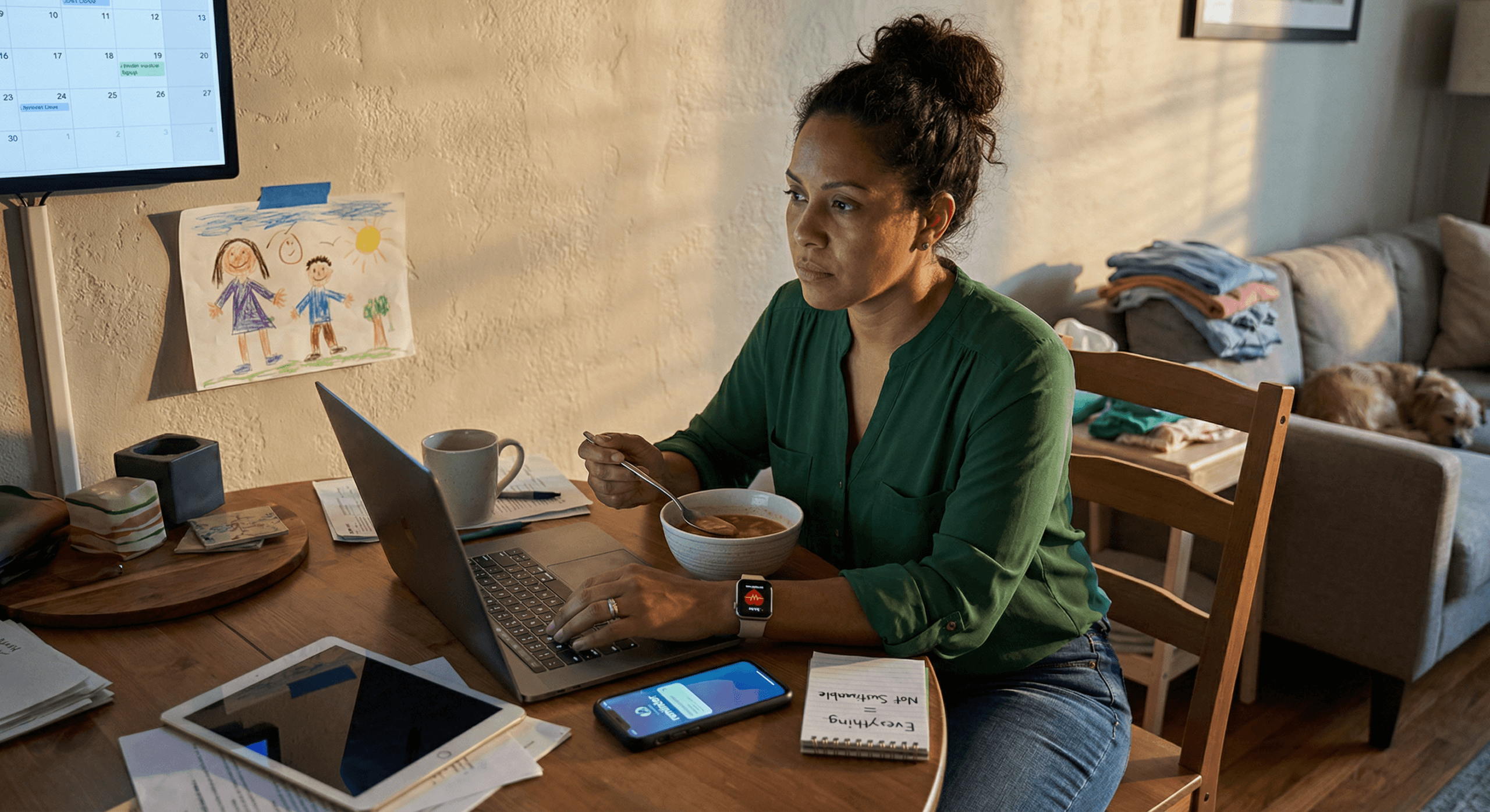 A woman pauses mid‑multitask at her home desk surrounded by devices and unfinished work, reflecting the shift toward delegating without guilt and releasing the pressure to do it all alone.